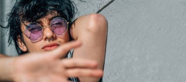 Eski gözlüklü genç adam duvara yaslanıyor ve elinin önünde duruyor.