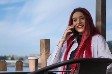 Genç kızıl saçlı kız dışarıda kahve içiyor ve cep telefonu kullanıyor.