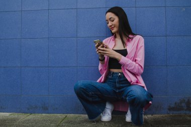 Gülümseyen genç kadın mavi duvarda cep telefonu kullanıyor.