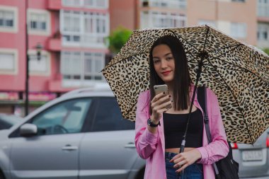 Yağmurda şemsiyeli genç bir kadın sokakta akıllı telefon kullanıyor.