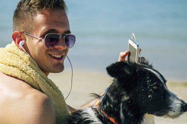 Kulaklıklı genç adam köpeğiyle sahilde eğleniyor.