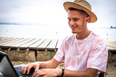 Kumsaldaki terasta dizüstü bilgisayar kullanarak rahat genç bir adam.