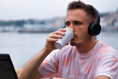 Plaj terasında dizüstü bilgisayar kullanan ve kahve içen, kulaklık takan rahat genç bir adam.