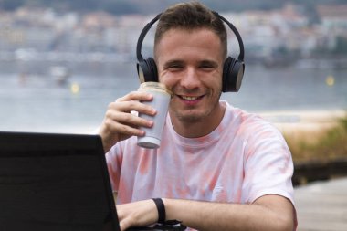 Plaj terasında dizüstü bilgisayar kullanan ve kahve içen, kulaklık takan rahat genç bir adam.