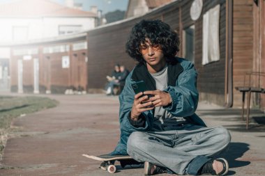 Kaykayının üstünde oturan genç adam cep telefonu kullanıyor.