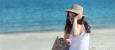 Şapkalı ve güneş gözlüklü kız sahilde oturup denize bakıyor.
