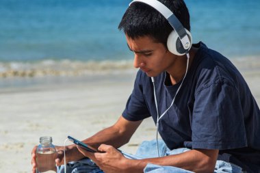 Kulaklıklı genç adam sahilde cep telefonuyla oynuyor.