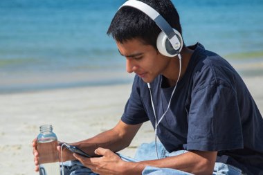 Kulaklıklı genç adam sahilde cep telefonuyla oynuyor.