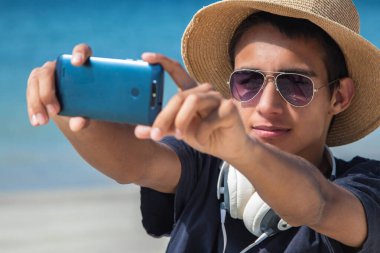 Şapkalı ve güneş gözlüklü genç adam sahilde telefonla selfie çekiyor.
