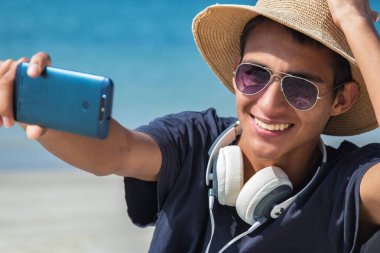 Şapkalı ve güneş gözlüklü genç Latin adam sahilde telefonla selfie çekiyor.