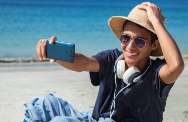 Genç adam sahilde telefonla selfie çekiyor.