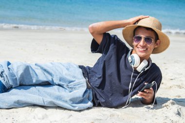 Plajda duran kulaklıklı ve cep telefonlu rahat genç bir adam.