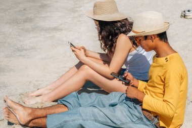 Plajda oturan gençler cep telefonu kullanıyorlar.