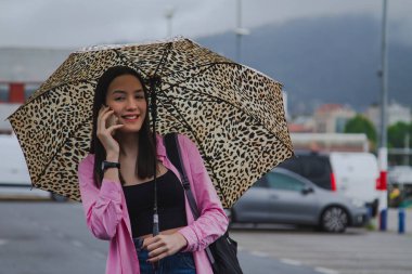 Gülümseyen genç kadın telefonda konuşuyor ve şemsiyesi yağmurda sokakta.