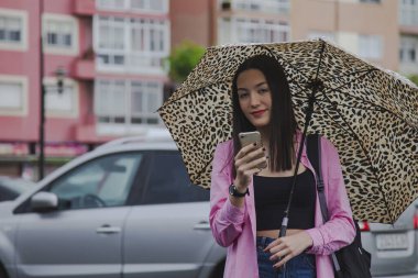 Yağmurda şemsiyeli genç bir kadın sokakta akıllı telefon kullanıyor.