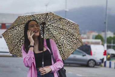 Gülümseyen genç kadın telefonda konuşuyor ve şemsiyesi yağmurda sokakta.