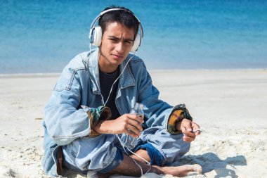 Latin kökenli genç adam plajda kulaklıklarla rahatlıyor ve içki içiyor.