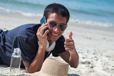 Sahilde cep telefonlu genç bir adam ve iyi bir jest yapıyor.
