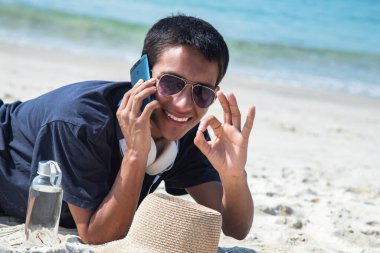 Sahilde cep telefonlu genç bir adam ve iyi bir jest yapıyor.