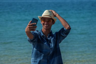 Genç adam sahilde telefonla selfie çekiyor.