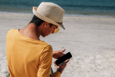 Latin asıllı genç adam plajda cep telefonuyla dinleniyor.