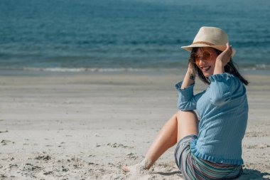 Güneş gözlüklü, şapkalı, kumsalda oturan mutlu kadın.