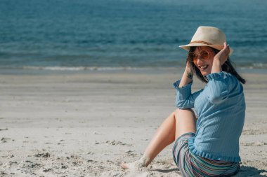 Güneş gözlüklü, şapkalı, kumsalda oturan mutlu kadın.