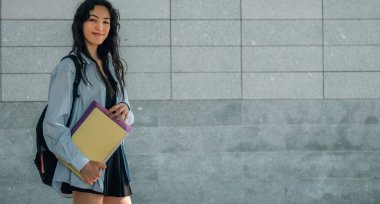 Sırt çantalı ve sokak duvarında kitapları olan öğrenci kızın portresi.