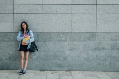 Gülümseyen öğrenci sokakta duvara yaslanıyor.