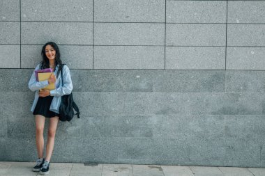 Sırt çantalı ve sokak duvarında kitapları olan öğrenci kızın portresi.