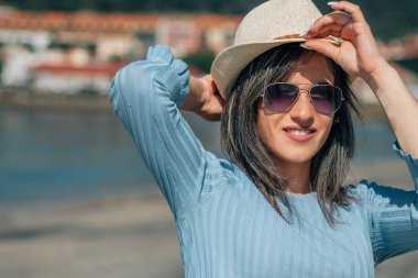 Sahilde dinlenen şapkalı gülümseyen genç bir kadının portresi.