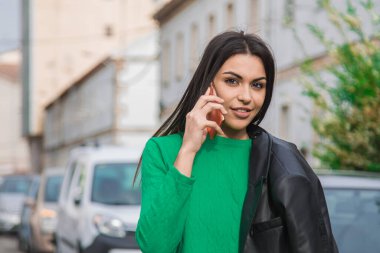 Deri ceketli şehirli kadın sokakta cep telefonuyla konuşuyor.