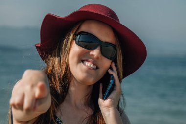 Kumsalda cep telefonuyla konuşan ve parmağıyla işaret eden kıvrımlı sarışın kız.