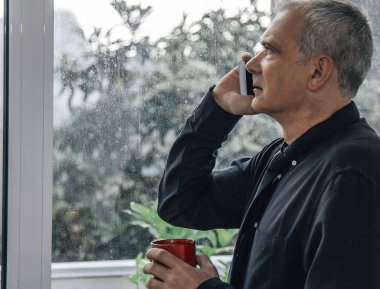 İş adamı pencereden dışarı bakıyor ve kahve içerken telefonla konuşuyor.