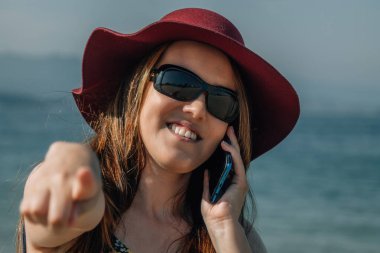 Kumsalda cep telefonuyla konuşan ve parmağıyla işaret eden kıvrımlı sarışın kız.