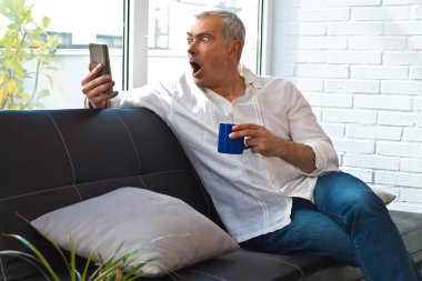 Şaşkın bir ifadeyle orta yaşlı bir adam telefona bakıyor ve kanepede kahve içiyor.