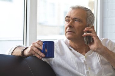 Olgun bir adam evinde kahve ve cep telefonuyla kanepede dinleniyor.