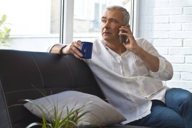 Olgun bir adam evinde kahve ve cep telefonuyla kanepede dinleniyor.