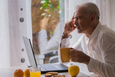 Yetişkin bir adam evde kahvaltı ederken dizüstü bilgisayarla çalışıyor.