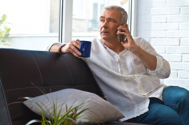 Olgun bir adam evinde kahve ve cep telefonuyla kanepede dinleniyor.