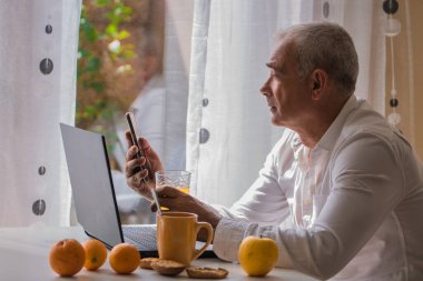 Orta yaşlı bir adam laptop ve cep telefonuyla çalışırken evde kahvaltı yapıyor.