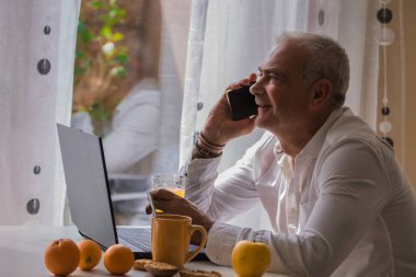 Orta yaşlı bir adam laptop ve cep telefonuyla çalışırken evde kahvaltı yapıyor.