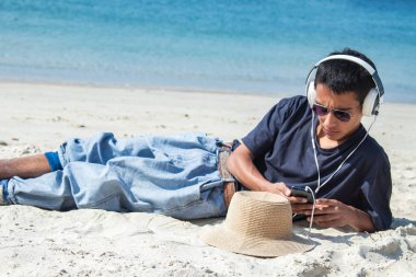 Sahilde kulaklığı ve telefonu olan genç bir adam.