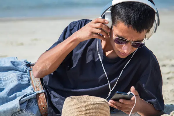 Sahilde kulaklığı ve telefonu olan genç bir adam.