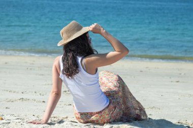 Genç bir kadın kumsalda oturmuş denizi düşünüyor.