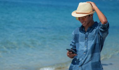 Genç adam sahilde yürürken cep telefonu kullanıyor.