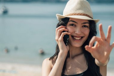 Kumsalda gülümseyen kadın telefonda konuşuyor ve parmaklarıyla işaret veriyor.