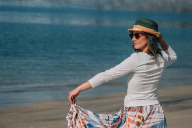 Yazın sahilde gülümseyen genç kadın moda modası kıyafetleri tasasız ve mutlu, özgürlüğü hisseden, boho tarzı şık ve güneş gözlüklü.