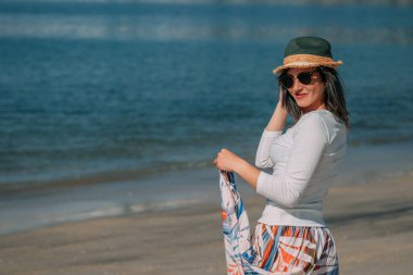 Yazın sahilde gülümseyen genç kadın moda modası kıyafetleri tasasız ve mutlu, özgürlüğü hisseden, boho tarzı şık ve güneş gözlüklü.