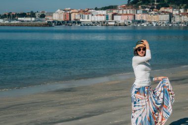 Yazın sahilde gülümseyen genç kadın moda modası kıyafetleri tasasız ve mutlu, özgürlüğü hisseden, boho tarzı şık ve güneş gözlüklü.